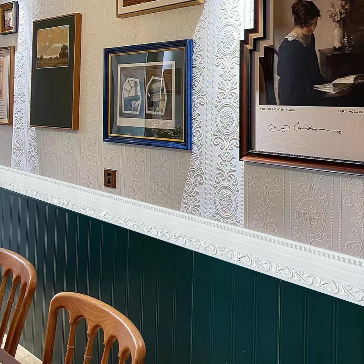 Framed artwork on a wall with decorative molding and wooden chairs below.